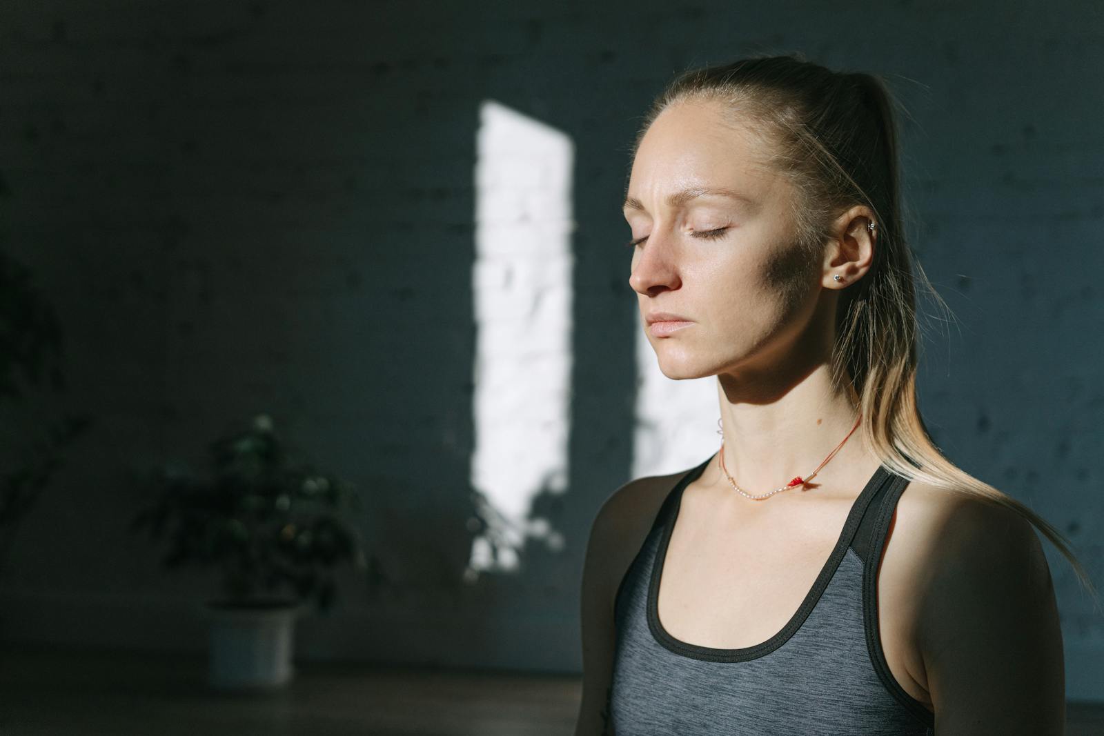 Woman with eyes closed, meditating in a sunlit room, wearing a gray athletic tank top.