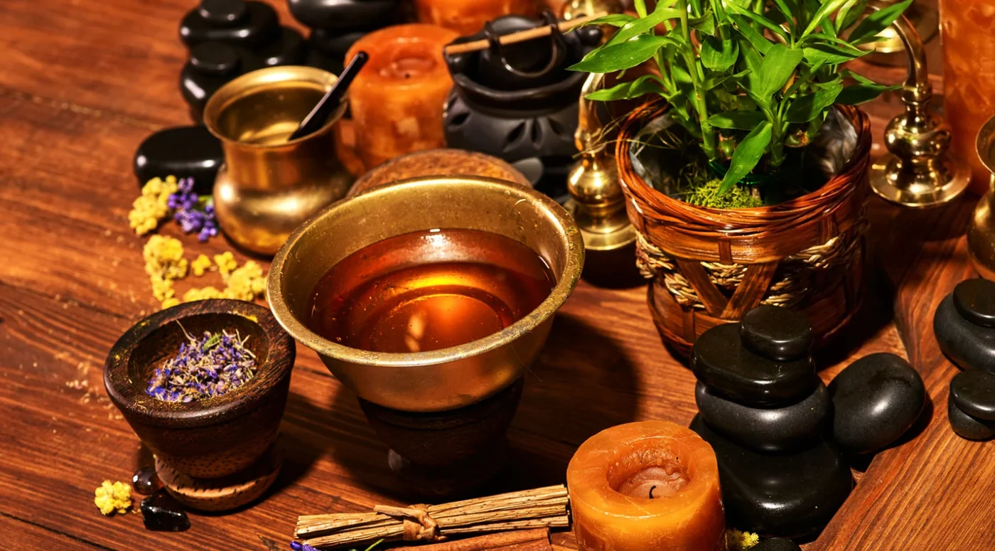 Relaxing spa scene with a copper bowl, dried herbs, candles, and a potted plant on a wooden table.