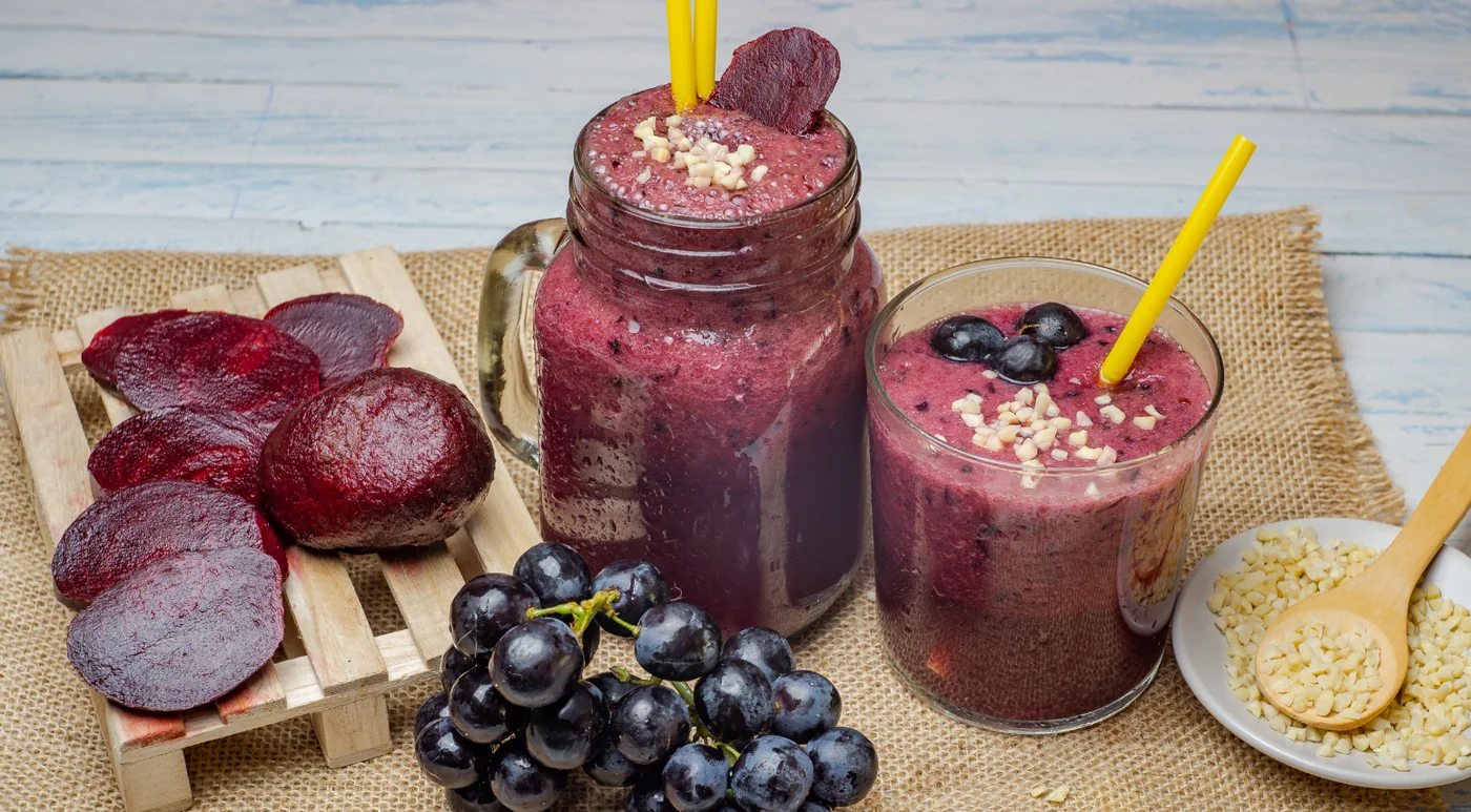 Two purple beet-grape smoothies in jars with yellow straws, surrounded by raw beets, grapes, and a plate of chopped almonds on a burlap surface