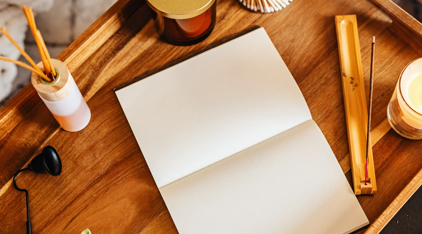 Open notebook with blank pages on a wooden desk, surrounded by a candle, incense, and writing tools on a tray.