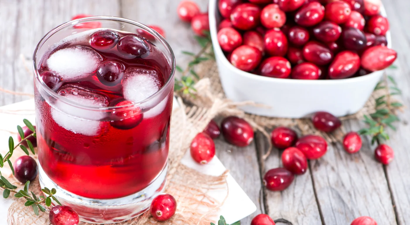 Glass of cranberry juice with ice and fresh cranberries on a rustic wooden table, nearby white bowl full of cranberries and greens for garnish