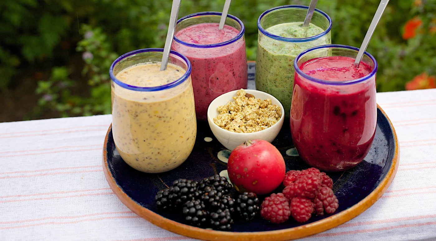 Tray of four smoothies in blue-rimmed glasses (yellow, pink, green, red) with berries, apple, and granola on a dark platter outdoors.