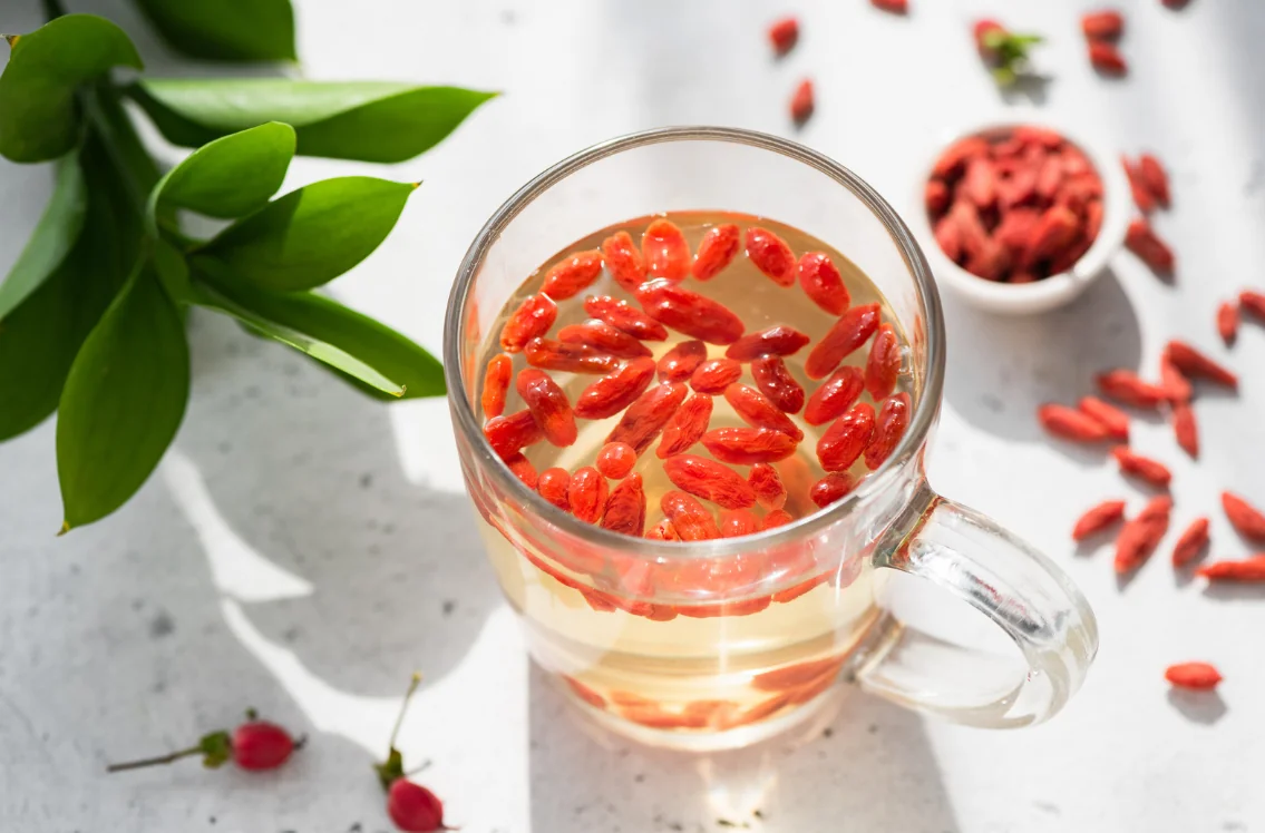 Glass mug of pale tea with red goji berries floating on the surface, scattered berries on a white surface with a small bowl nearby and green leaves in the background.