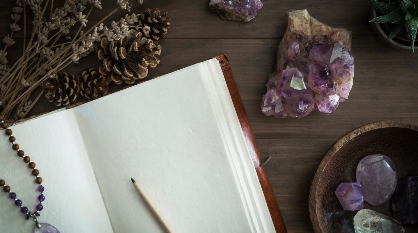 Open notebook with a pencil, beaded necklace, pinecones, and purple crystals on a wooden desk.