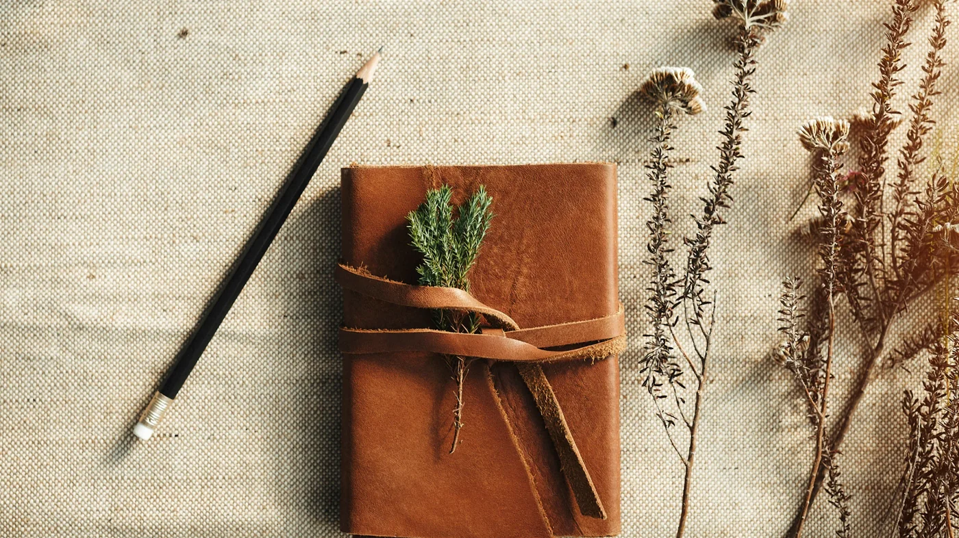 Brown leather journal tied with a strap, a sprig tucked inside, on burlap with a pencil nearby.