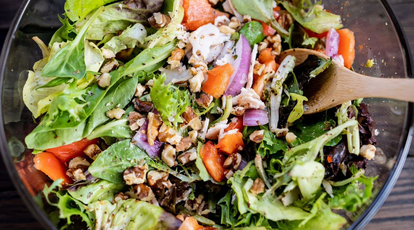 Bowl of mixed greens salad with lettuce, carrot chunks, red onion, and chopped walnuts with a wooden spoon resting inside