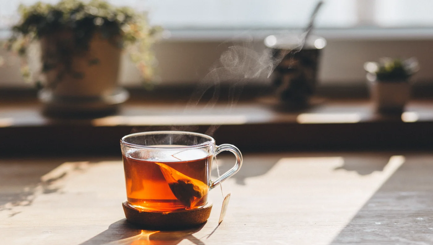 Glass cup of hot tea steaming on a wooden table near a sunlit windowsill with a tea bag tag visible beside it.