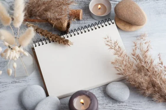 Blank spiral notebook on a pale wooden surface, surrounded by dried plants, candles, and smooth stones.