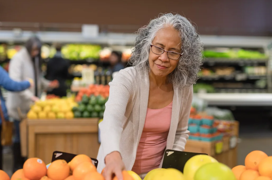 Older woman with gray curly hair and glasses selecting oranges in a grocery store produce section.