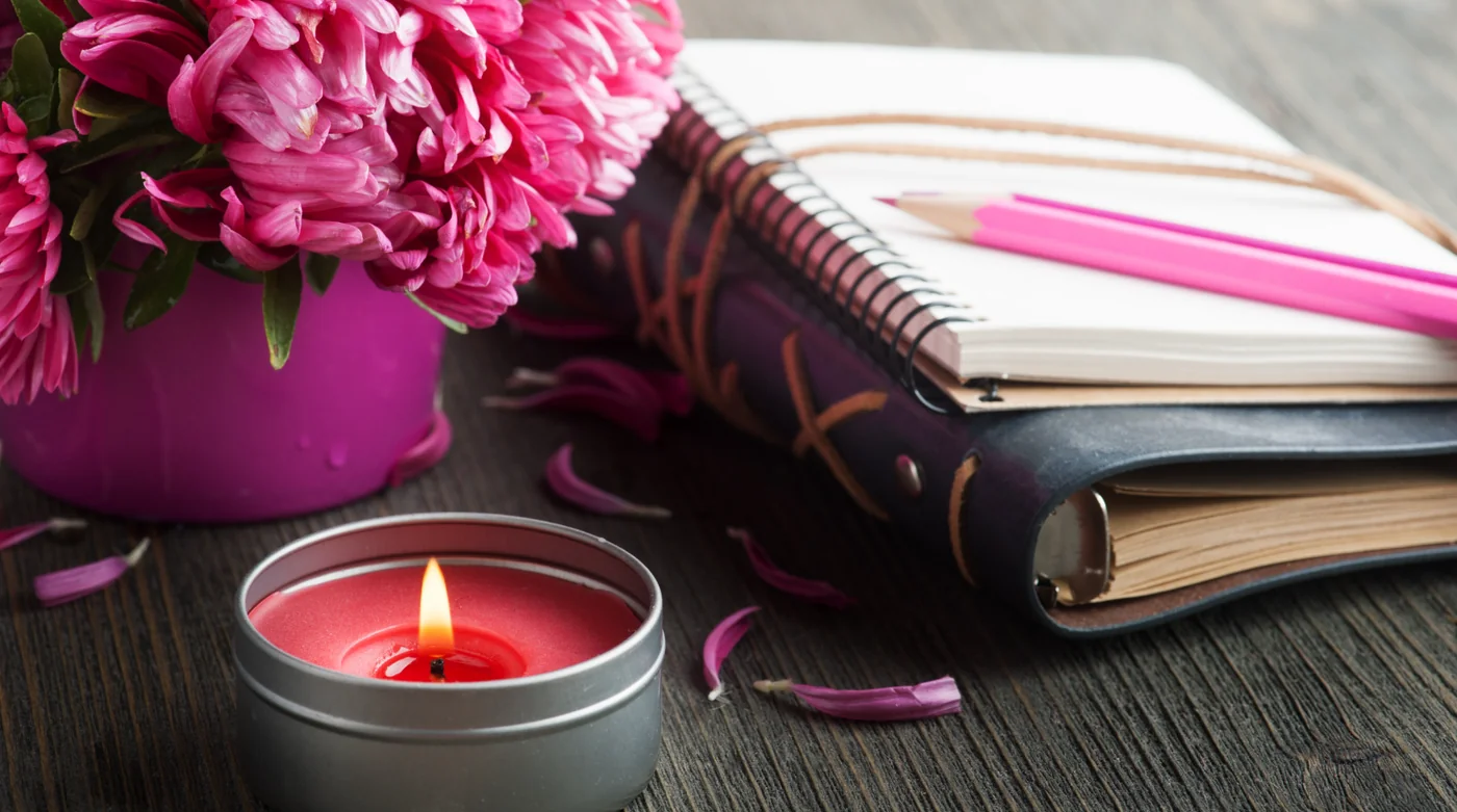 Pink bouquet in a pink pot beside a spiral notebook and pink pencils on a dark wooden desk with a lit candle nearby.