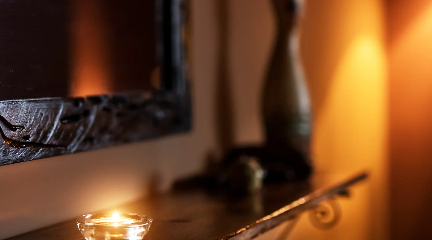 Small lit tealight on a wooden mantel, with a blurred bottle in the warm background creating cozy ambience.