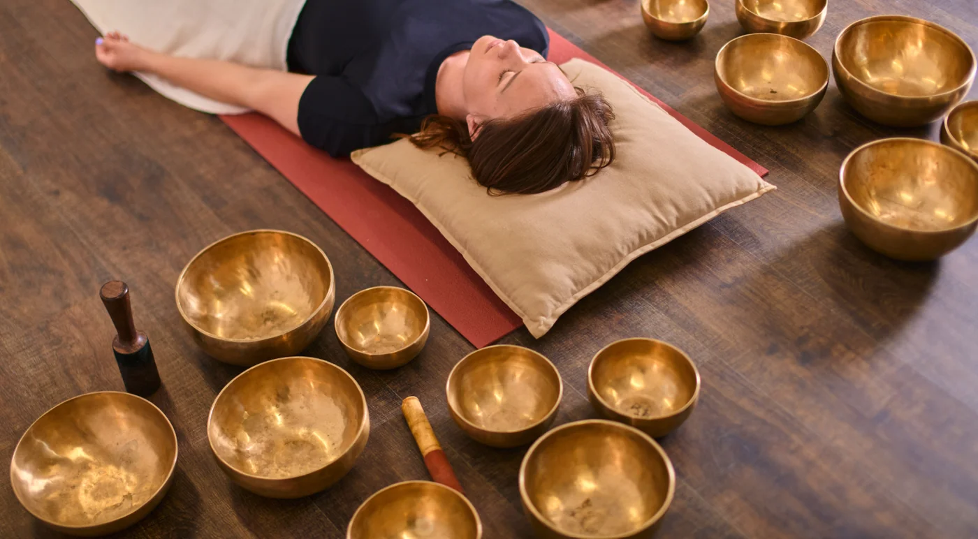 Woman lying on a pillow on a red mat among brass singing bowls on a wooden floor, ready for a sound bath therapy session.