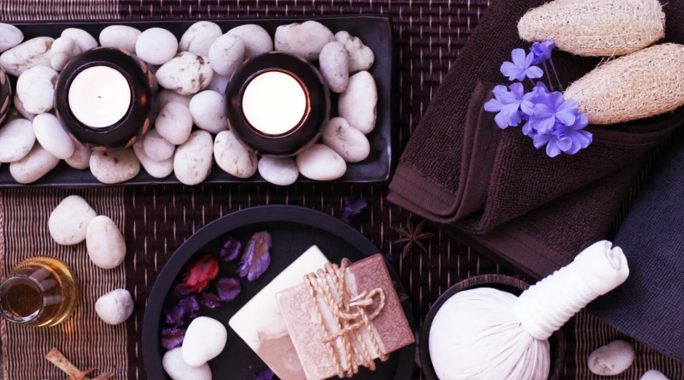 Spa flat lay with candles in dark bowls, white pebbles, brown towels, a purple flower, soaps, oil, and wrapped gifts on a mat.