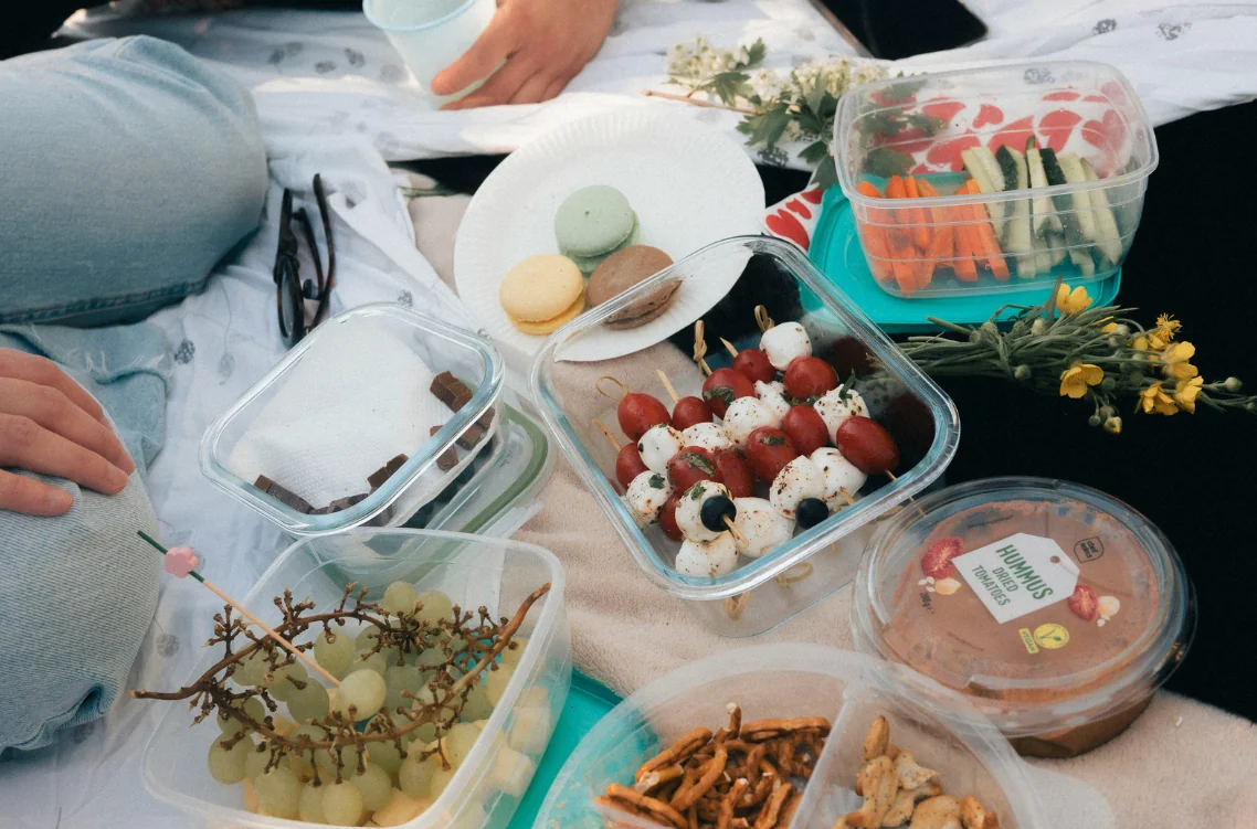 Picnic spread on a blanket with clear containers of grape skewers, macarons, hummus, and veggie sticks nearby.