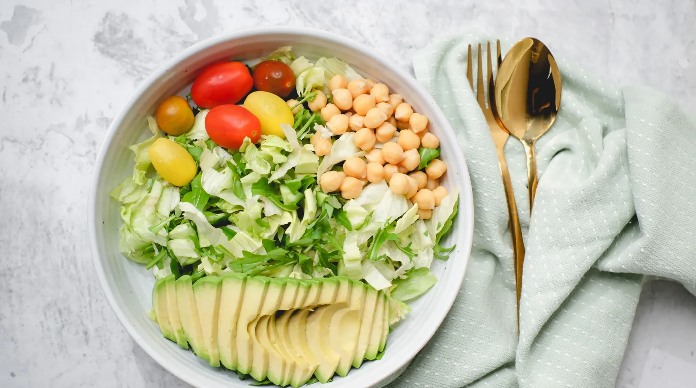 Bowl of salad with mixed greens, cherry tomatoes, chickpeas, and sliced avocado with gold cutlery on a pale napkin.