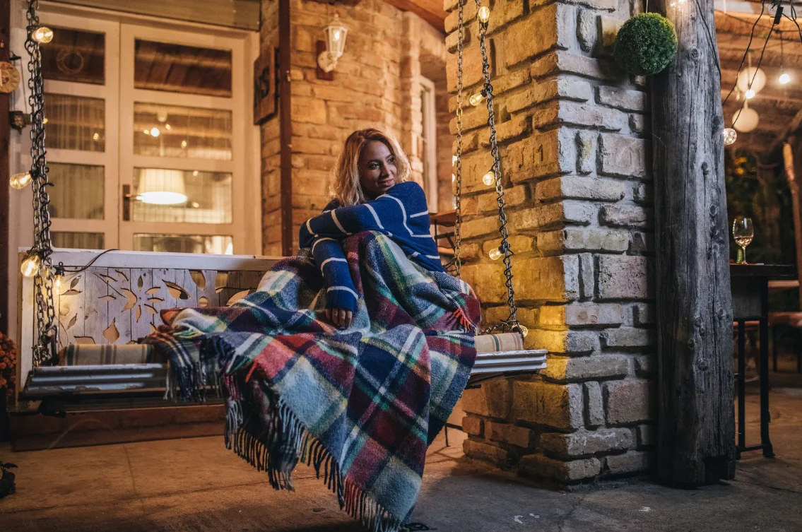 Woman on a hanging porch swing, wrapped in a blue striped sweater and a plaid blanket, with warm string lights nearby.